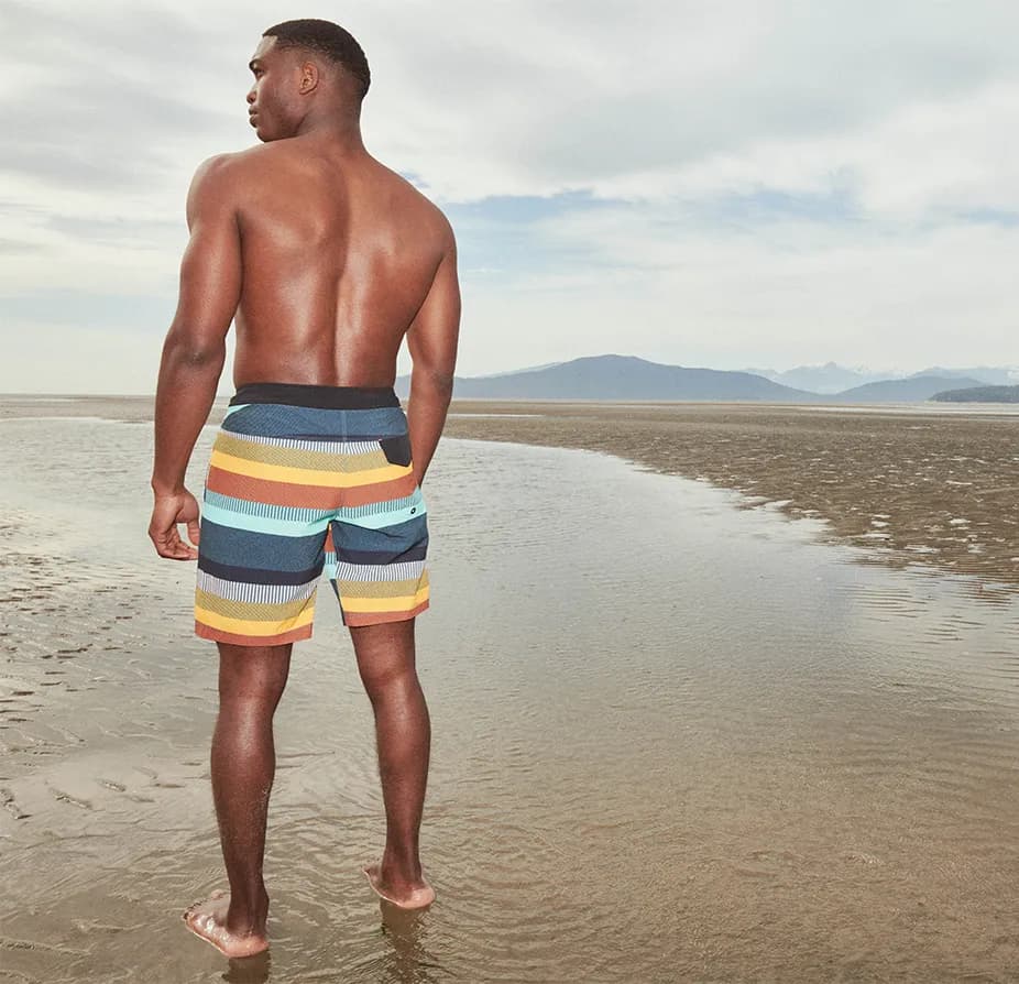 A man wearing swim trunks stands confidently on the beach, with the ocean waves in the background and the sun shining brightly