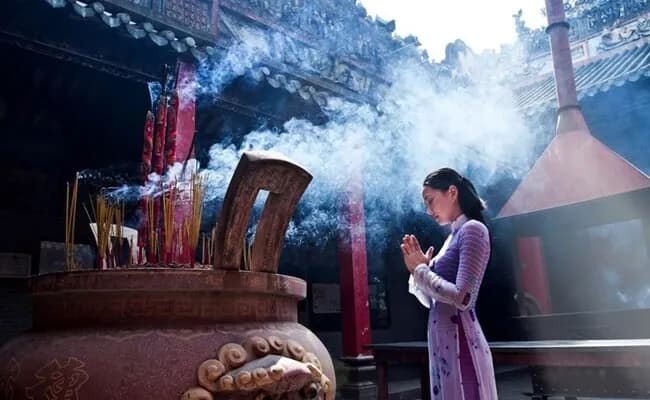 A Vietnamese woman elegantly dressed in a colorful ao dai stands inside a temple, surrounded by traditional architecture and peaceful decorations