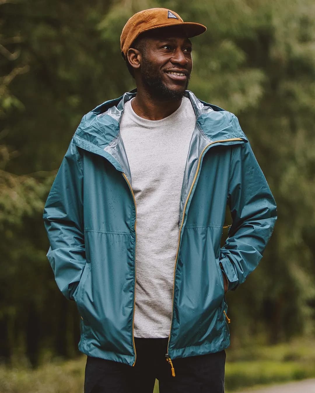 A man wearing a lightweight jacket over a t-shirt and a cap stands casually outdoors, exuding a relaxed vibe against a scenic backdrop