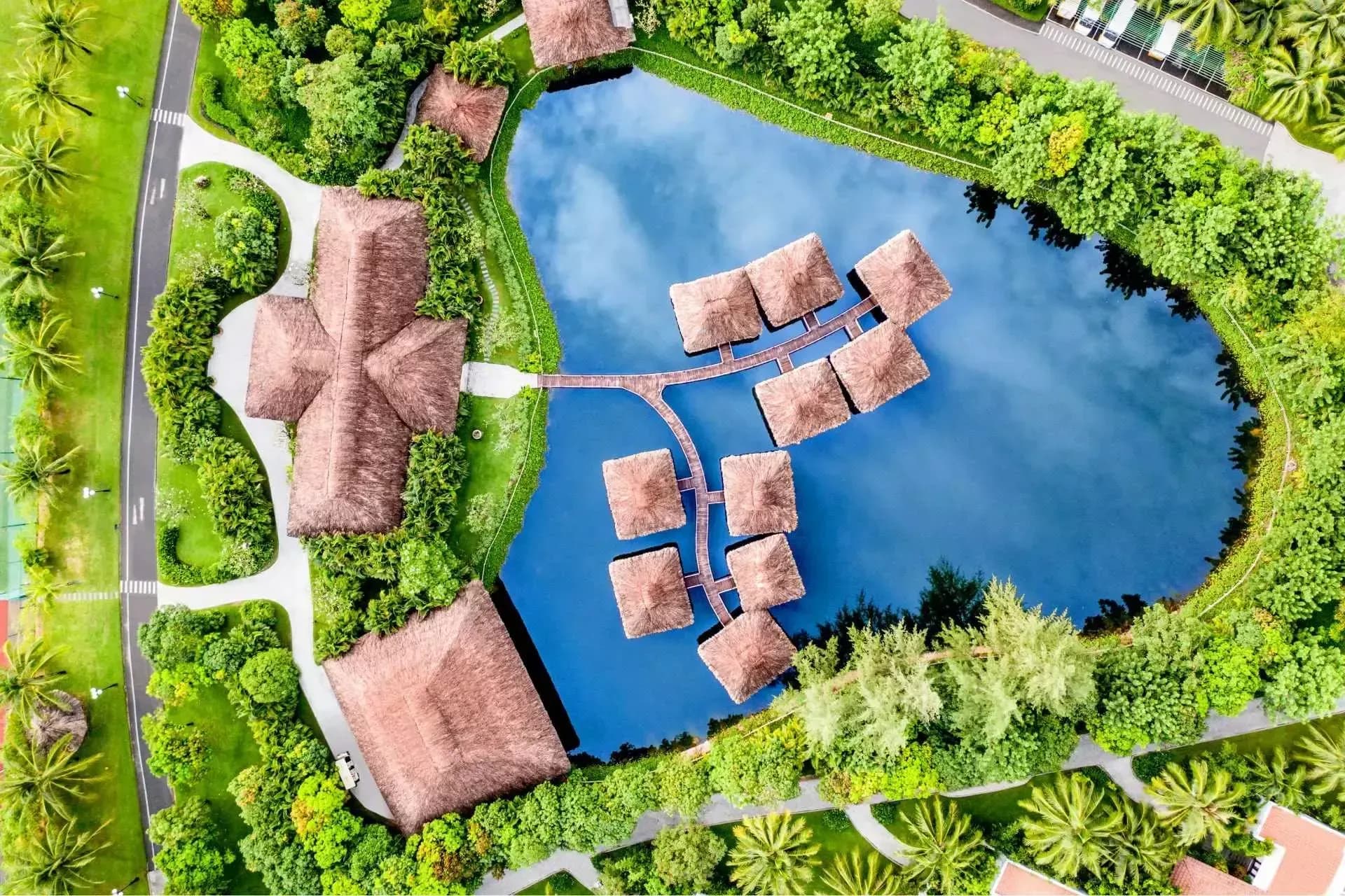 Resort area with thatched roofs and a blue lake viewed from above, surrounded by greenery and walking paths.